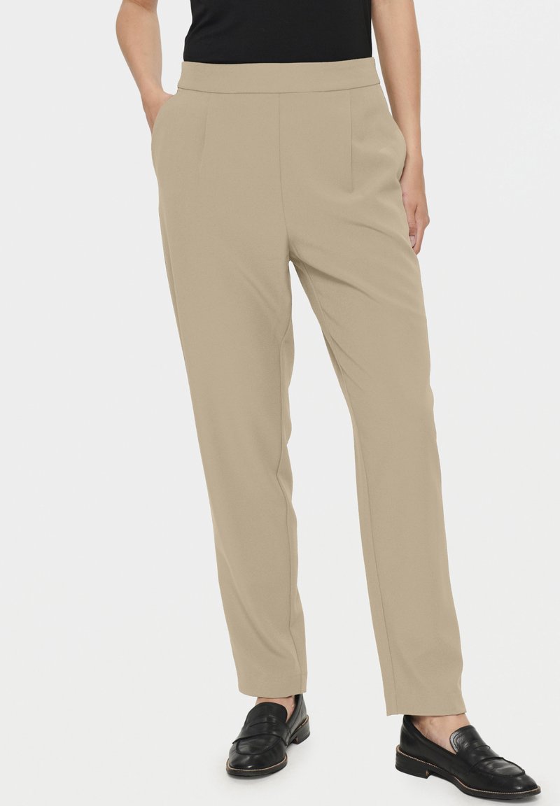 Pantalons beige ajustés au toucher doux, avec des poches latérales et une coupe droite, associés à des mocassins noirs.