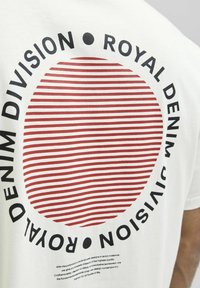 Hvid bomulds T-shirt med et cirkulært rødt stribet grafisk design, der har sort tekst, der læser "ROYAL DENIM DIVISION" rundt om.