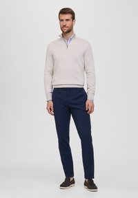 Maglione zip-neck beige chiaro, pantaloni blu navy e scarpe slip-on scure. L'outfit presenta un design semplice e colori uni con pantaloni risvoltati.
