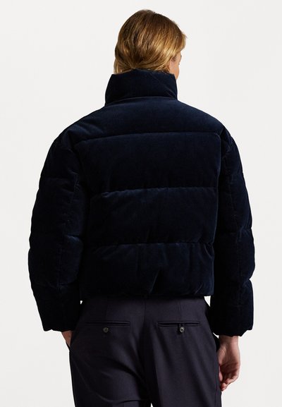 Marineblauwe corduroy pufferjack met een hoge kraag en een horizontaal geborduurd ontwerp. Beschikt over een ontspannen pasvorm en brede mouwen.