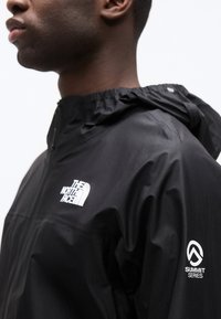 Μαύρο μπουφάν με κουκούλα, με λευκά λογότυπα The North Face και Summit Series, φορεμένο από άτομο που κοιτάζει αριστερά σε λευκό φόντο.