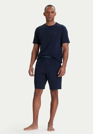 Modello maschile che indossa una camicia blu navy a maniche corte e pantaloncini abbinati, in piedi a piedi nudi su uno sfondo semplice.