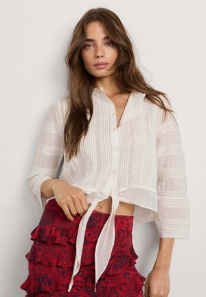 Jonge vrouw met lang bruin haar draagt een doorschijnende witte blouse met knopen, gestrikt in de taille, en een rode, met patroon versierde, gelaagde rok.