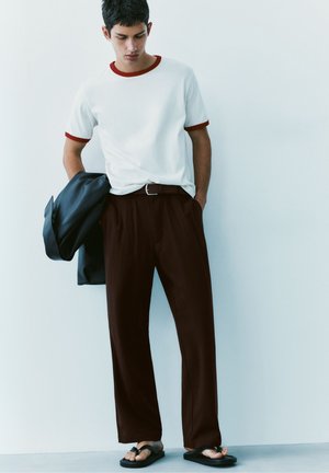 Jeune homme en t-shirt blanc avec bordure rouge, pantalon marron, tongs noires, tenant une veste sombre, debout contre un mur blanc.
