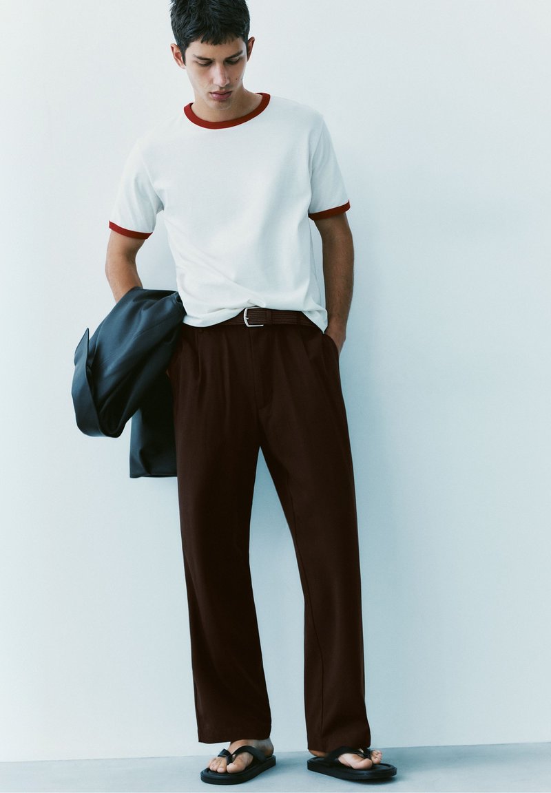 Jeune homme en t-shirt blanc avec bordure rouge, pantalon marron, tongs noires, tenant une veste sombre, debout contre un mur blanc.