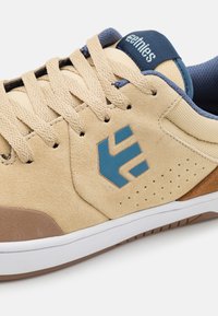 Beige velúr sportcipő kék logóval, texturált részletekkel és barna sarokkal. Kerek orrú, szabványos fűzéssel és fehér gumitalppal rendelkezik.