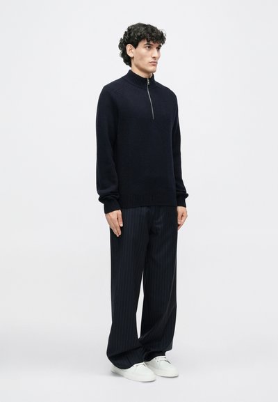 Dark navy pullover sweater with a high collar and a partial zipper, worn with pinstriped trousers and white sneakers. Soft knit texture.