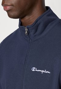 Μπλε σακάκι με φερμουάρ και ψηλό γιακά, κατασκευασμένο από μαλακό ύφασμα. Διαθέτει λευκό κεντημένο λογότυπο στο στήθος και λείο φερμουάρ.