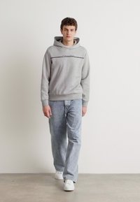 Grå hettegenser med et frontlogo, kombinert med lyseblå rette jeans og hvite sneakers mot en nøytral bakgrunn.