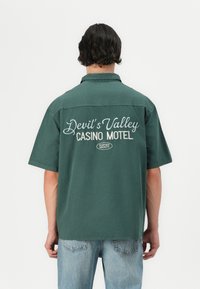 Rövid ujjú zöld póló, pamutból készült, fehér hímzett felirattal a hátsó részén: "Devil's Valley CASINO MOTEL". Kényelmes szabású és sima tapintású.
