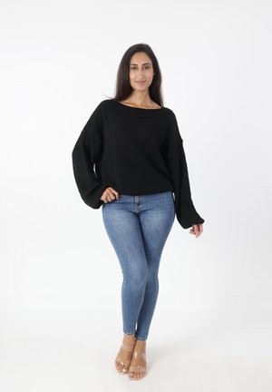 Zwarte gebreide trui met een ruime halslijn en lange, klokvormige mouwen, gecombineerd met aansluitende blauwe jeans en doorzichtige hakken.