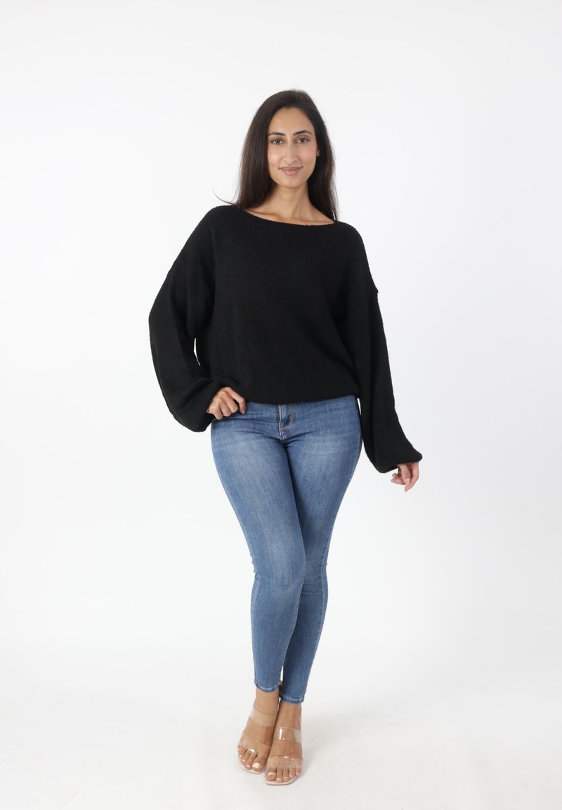 Zwarte gebreide trui met een ruime halslijn en lange, klokvormige mouwen, gecombineerd met aansluitende blauwe jeans en doorzichtige hakken.