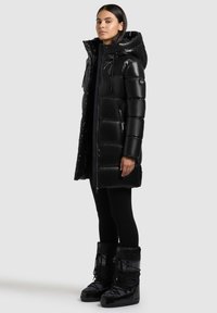 Sort puffy parka med hætte, blank finish, lynlåslukning foran og sidelommer. Båret med sorte leggings og matchende højhælede støvler.