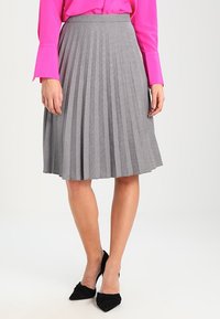 Šedá kostičkovaná plisé sukně s přiléhavým pasem, do délky kolen, vyrobená z lehkého materiálu, nošená s černými lodičkami na vysokém podpatku.