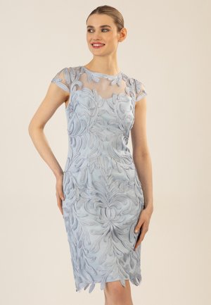 Frau trägt ein ärmelloses, knielanges hellblaues Kleid mit kunstvoll bestickten Blumenmustern, lächelt, mit zurückgebundenem Haar.