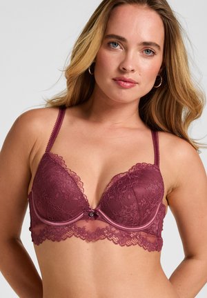 Woman with long blonde hair wearing a plum-colored lace bra with thin straps against a plain light background.