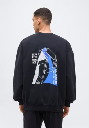 Mann von hinten, trägt schwarzen Pullover mit geometrischem weißen und blauen Design und dem Text "Peak Performance" sowie "Find the flow see where free takes you."