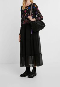 Langes schwarzes Kleid mit floralem Stickerei, durchsichtigen langen Ärmeln und gestuftem Rock. Kombiniert mit schwarzen strukturierten Knöchelstiefeln und einer schwarzen Tasche.