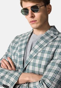 Marynarka w kratę w odcieniach szarości z zielonymi i niebieskimi akcentami, z wykładanym kołnierzem i klasycznym krojem. W połączeniu z ciemnymi, okrągłymi okularami przeciwsłonecznymi.