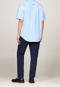 Hellblaues kurzärmliges Hemd mit abgerundetem Saum, Logodetail am Kragen, kombiniert mit marineblauen Anzughosen und schwarzem Schuhwerk.