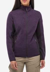 Femme portant une veste polaire zippée violette avec des panneaux latéraux noirs et des poches zippées, associée à un pantalon beige.