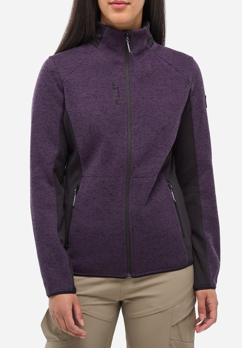 Femme portant une veste polaire zippée violette avec des panneaux latéraux noirs et des poches zippées, associée à un pantalon beige.