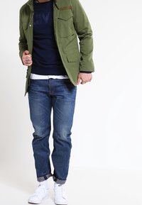 Chaqueta verde oliva con múltiples bolsillos, suéter azul marino debajo, jeans de color azul claro y zapatillas blancas. Atuendo casual en capas.