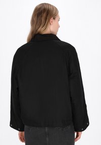 Giacca nera con colletto in velluto a coste, vestibilità rilassata e dettagli con bottoni a pressione sulle maniche. La vista posteriore mette in risalto la texture liscia e i dettagli delle cuciture.