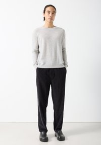 Muškarac stoji s rukama u džepovima, nosi svijetlosivi džemper, crne somotske hlače i crne cipele bez vezica na bijeloj pozadini.