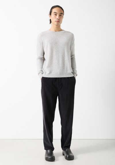 Homme debout les mains dans les poches, portant un pull gris clair, un pantalon en velours côtelé noir et des chaussures noires sans lacets, sur fond blanc.