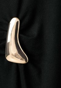 Accent métallique doré avec une forme lisse et courbée sur un tissu noir, créant des plis et de la texture. Le design présente une surface brillante et réfléchissante.