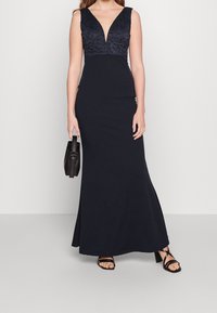 Marinblå klänning med djup V-ringning, åtsittande midja och utställd kjol. Har spetsdetaljer upptill och en mjuk tygstruktur.