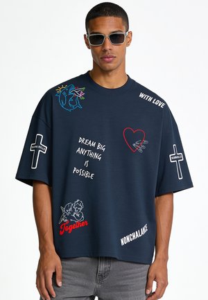 Mann trägt dunkelblaues oversized T-Shirt mit aufgedrucktem Kreuz, Hai, Herz, Engeln und Text, der "Dream Big," "Together," und "With Love." enthält