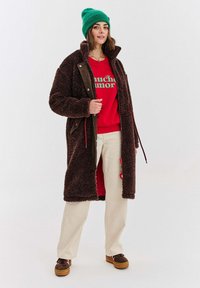 Žena v zelené čepici, červené mikině s textem, dlouhém hnědém koženém kabátu, bílých kalhotách a hnědých botách stojící proti jednobarevnému pozadí.