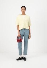 Gul strikket cardigan med korte ermer, blå rett ben jeans, svarte loafers, hvite sokker og en liten rød håndveske med detaljer i gull.