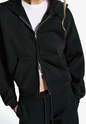 Person trägt schwarzen Kapuzenpullover mit Reißverschluss und Kängurutasche sowie passende schwarze Hose, weißes Hemd darunter sichtbar, Hände in den Taschen.