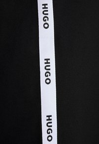 Tissu noir avec une bande blanche verticale comportant le mot Hugo répété en texte noir le long de la bande.
