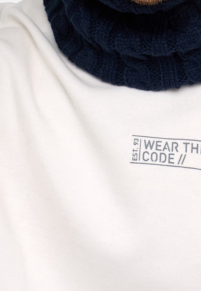 Tissu blanc avec le texte imprimé "EST. 93 WEAR THE CODE //" partiellement couvert par une écharpe en tricot bleu foncé.