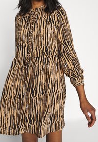 Ruskea lyhythihainen mekko, jossa on pystysuuntaiset mustat raidat, nappikiinnitteinen yläosa, gathered vyötärö ja pehmeä kankaan rakenne.