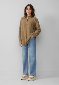 Světle hnědá button-up košile s texturovanou látkou, kombinovaná se světle modrými širokými džínami a krémovými platformovými sandály. Jednoduchý design a uvolněný střih.