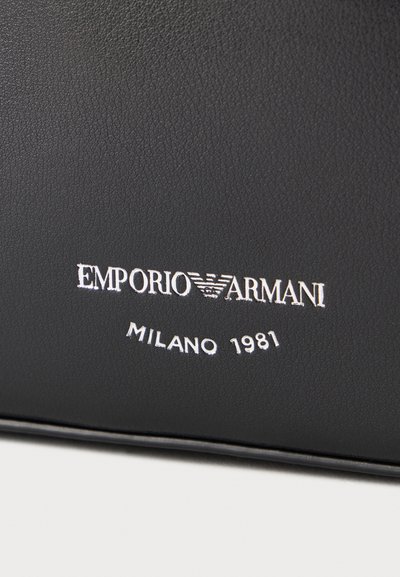 Μαύρη δερμάτινη επιφάνεια με λευκό λογότυπο Emporio Armani και το κείμενο «Milano 1981» ανάγλυφα κάτω από το όνομα της μάρκας.