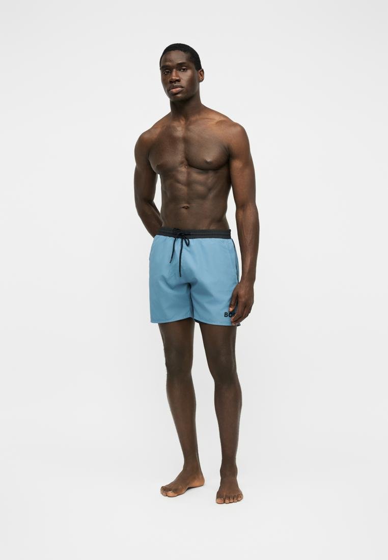 Homme musclé debout pieds nus portant un short de bain bleu clair avec une ceinture noire et un cordon sur un fond blanc uni.