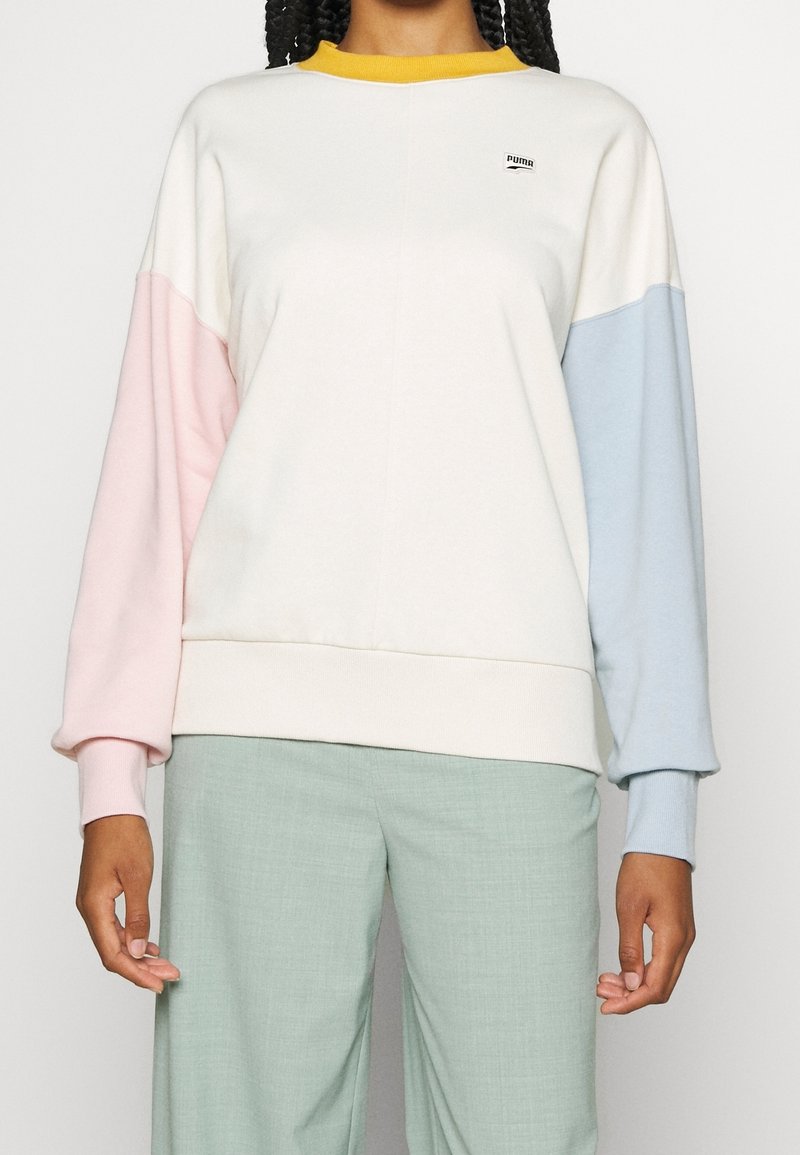 Personne portant un sweat-shirt blanc avec une manche gauche rose, une manche droite bleue et un col jaune, associé à un pantalon vert clair.