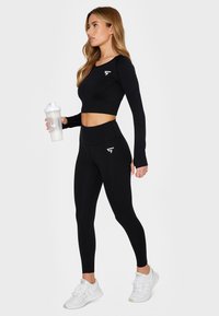 Black long-sleeve crop top and high-waisted leggings set. Features a fitted design, textured sleeves, and a white logo detail. Includes a shaker bottle.