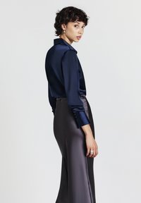 Donna con capelli corti e ricci si volta verso l'indietro, indossando una blusa di raso blu navy e una gonna di raso grigio scuro su uno sfondo neutro.