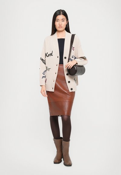 Cardigan beige con motivo testuale, top nero, gonna a matita in pelle marrone, collant neri e stivali al ginocchio marroni; borsa rotonda nera in mano.