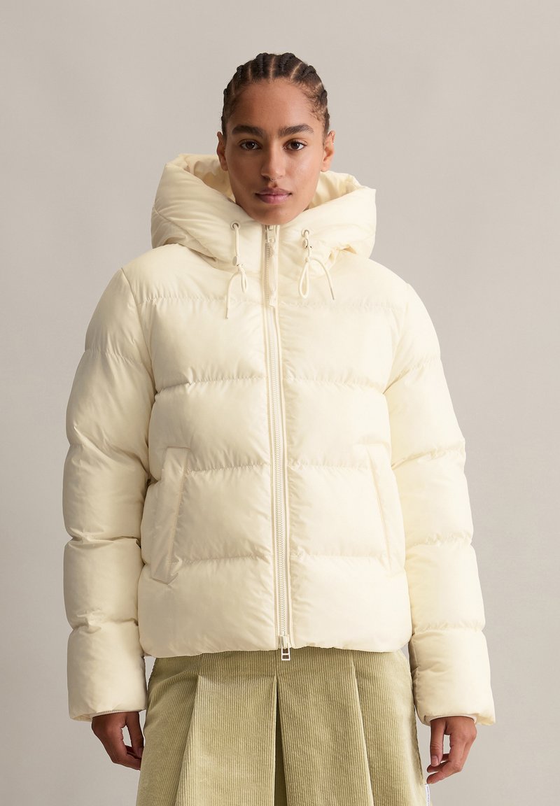 Pufferjacke in hellem Gelb mit gemütlicher Kapuze, Reißverschluss vorne und seitlichen Taschen. Mit gestepptem Muster und boxy Silhouette.