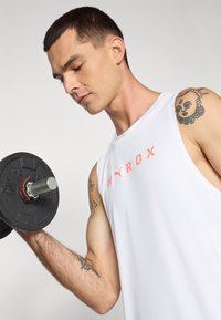 White sleeveless tank top with orange "HYROX" lettering, paired with a black weight plate in a solid grip, showcasing a muscular arm with tattoos.