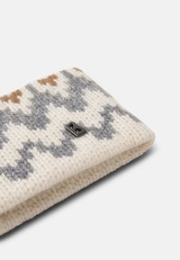 Strikket pandebånd i creme med zigzag-mønster i grå, brun og hvid. Har et lille metallisk logo-accents. Blødt, tekstureret stof.