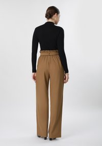 Femme aux cheveux foncés attachés en chignon, portant un haut noir à manches longues et un pantalon marron ample à taille haute, debout de dos.